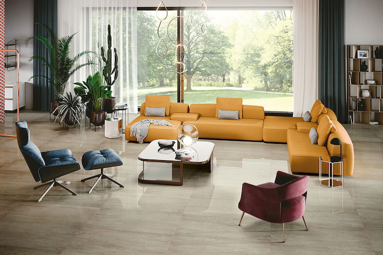Blick auf ein großzügiges Wohnzimmer mit modernen Bodenfliesen, einem riesigen gelben Sofa mit weiteren Sitzgelegenheiten sowie Pflanzen und einem Regal im Hintergrund und sehr großes bodentiefes Fenster mit Blick ins Grüne