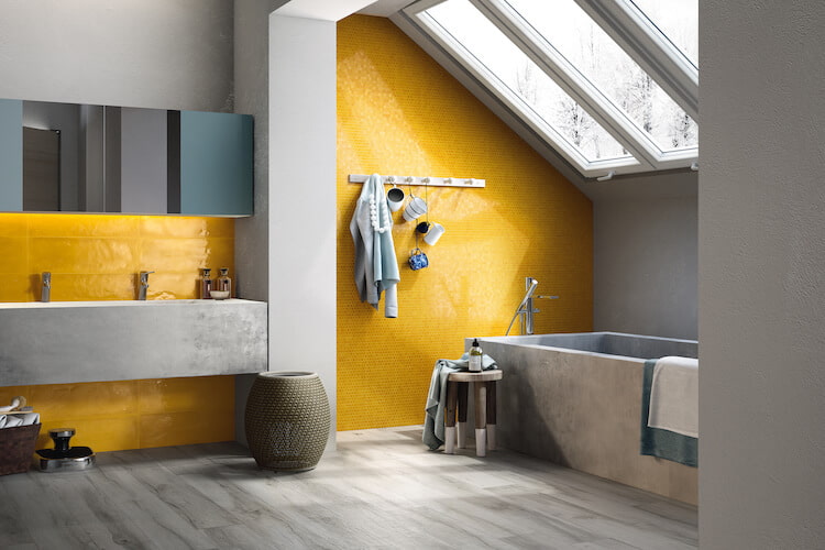 Kräftige gelbe Fliesen in einem sonst grau gehaltenden Badezimmer mit Blick auf Waschbereich, Badewanne und große, schräge Dachfenster