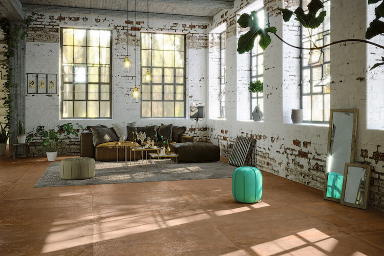 Terracotta-Bodenfliesen in einem Raum in Fabrikoptik mit großen Fenstern, abgenutzter Wand und Sitzgelegenheiten